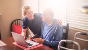 A woman leans over the shoulder of an elderly man seated before a computer. They smile at one another as she teaches him.